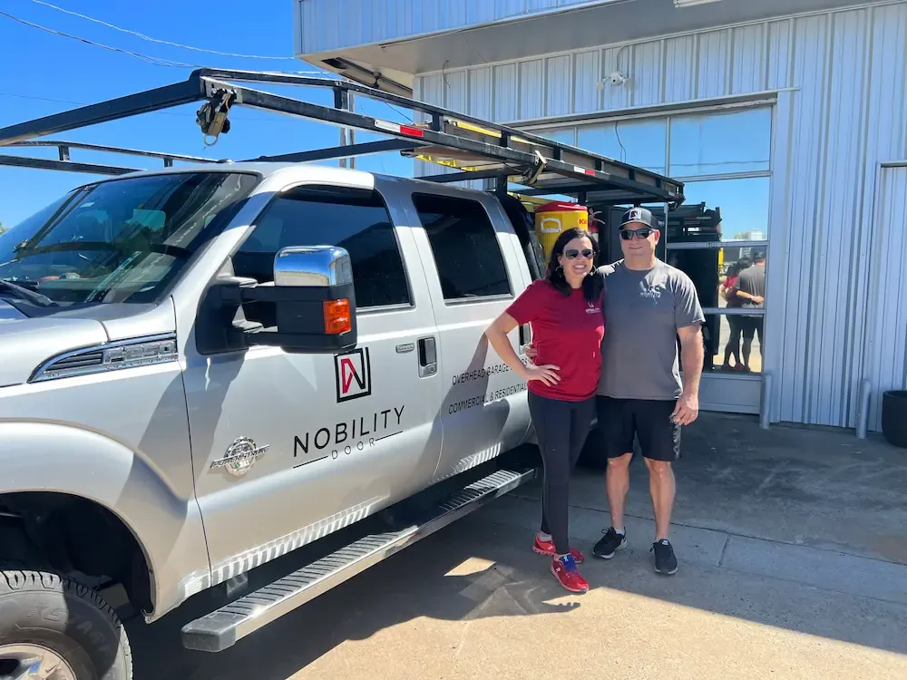 A man and woman standing in front of a silver 'Nobility Door' truck, smiling and posing near a building with a large window.